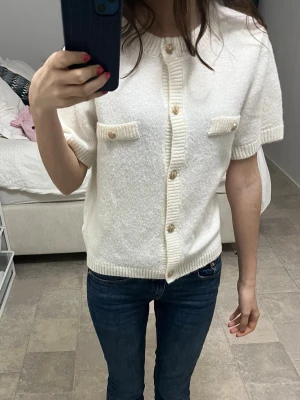 Vit kortärmad kofta Gina Tricot - Kortärmad kofta i mjukt, stickat material från Gina Tricot. Färgen är krämvit och koftan har dekorativa guldfärgade knappar framtill samt två fejkfickor på bröstet. Ribbstickade kanter vid ärmslut, hals och nederkant för en clean look.