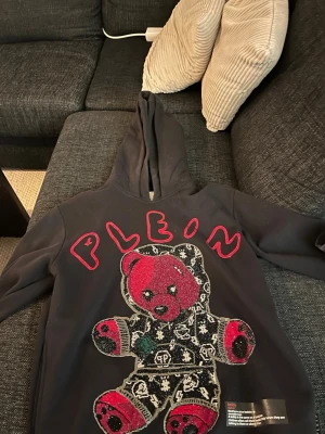 Svart hoodie Plein med nallemotiv storlek s - Svart hoodie från Plein med stor glittrig nalleapplikation på framsidan och röd text. Hoodien har huva och coola detaljer i både svart, rött och silver. Perfekt för dig som vill sticka ut med en unik streetstyle-look. Nästan aldrig använd fåtal gånger, köpt 2022 på deras hemsida för 6500kr med 40% rabatt, nästan bara hängt på en galje sedan dess, mail kvittot kan fås med vid köp.