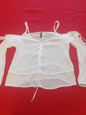 Vit transparent blus med knappar - Söt vit blus från Divided med transparent tyg, tunna axelband och off shoulder-långa ärmar. Blusen har knappar framtill och en dragsko i midjan för justerbar passform. Perfekt för en trendig och luftig look.