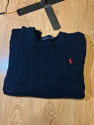 Mörkblå kabelstickad tröja Polo Ralph Lauren - Snygg mörkblå kabelstickad tröja från Polo Ralph Lauren. Tröjan har rund halsringning och ribbstickade muddar vid ärmslut och nederkant. Perfekt för dig som gillar klassisk och stilren design. Finns väldigt små defekter på halskragen.