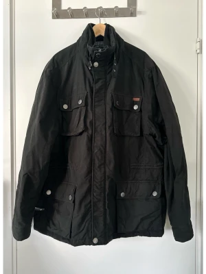 Svart Hampton Republic jacka, XXL - Snygg svart jacka från Hampton Republic i storlek XXL med stiligt rutigt foder. Jackan har flera fickor framtill med knappar och en innerficka med dragkedja. Kragen kan stängas högt upp med knappar. Ärmsluten har justerbara knappar. Intern dragsko i midjan för bästa passform. Fint skick!!  Hämtas i Stockholm eller skickas mot porto. 