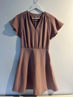 Rosa klänning  - Kortklänning i rosa med v-ringad hals och korta ärmar. Klänningen har en markerad midja och lätt utställd kjol. Storlek: 34 