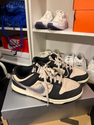 Nike Dunk Low svart/beige sneakers - Nike Dunk Low sneakers i svart och beige med silverfärgad Swoosh. Ovandelen är i läder och textil, med klassisk perforerad tåbox och svart yttersula. Skorna har beige snörning och låg silhuett som ger en clean och stilren look.