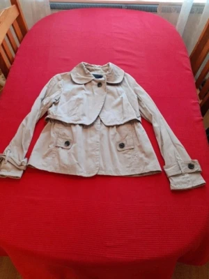 Kort beige trenchcoat med stora knappar - Snygg kort trenchcoat i beige med stora knappar och klassisk krage. Jackan har två fickor framtill med lock och knappar samt detaljerade ärmslut. Perfekt för dig som gillar stilrena och tidlösa plagg. Materialet känns lätt och passar bra till lager-på-lager.