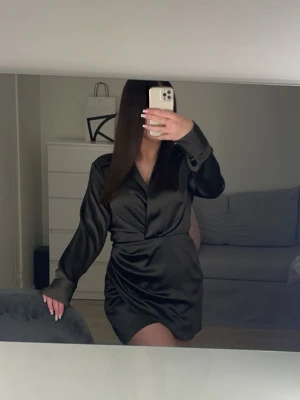 Satin klänning - En jättesnygg olivgrön kort omlottklänning i satin med v-ringning och långa ärmar. Klänningen har en lätt glansig finish och markerad midja som ger en elegant siluett. Perfekt för dig som vill ha en elegant och trendig look. Klänningen har en dold dragkedja vid midjan, för att kunna få på sig den på enklaste sätt.