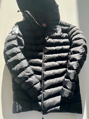 Svart dunjacka från U.S. Polo Assn. - Snygg svart dunjacka från U.S. Polo Assn. med huva och dragkedja framtill. Jackan har quiltad design, två sidofickor och logga på bröstet. Perfekt för kalla vinterdagar och riktigt skön att bära. Går att prata pris