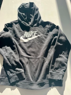 Svart Nike hoodie med stor logga - Säljer en svart hoodie från Nike med stor vit logga och swoosh på bröstet. Hoodien har en klassisk känguruficka framtill och huva. Tillverkad i mjukt bomullsmaterial som är skönt att ha på sig. Perfekt för dig som gillar sportig och avslappnad stil. Använt väldigt få gånger väldigt bra skick.