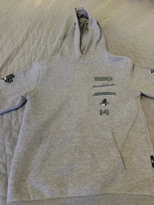 Grå hoodie från Hoodrich - Snygg grå hoodie från Hoodrich med tryck på bröstet och ryggen. Hoodien har huva, känguruficka och coola detaljer på ärmarna. Materialet är mjukt och skönt, perfekt för en avslappnad streetwear-look. Stora loggor och grafiska prints ger en modern vibe.