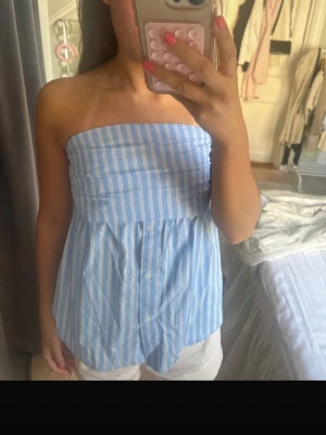 Blåvit randig bandeau tubtopp - Snygg bandeau tubtopp i blått och vitt med vertikala ränder och knappar framtill. Toppen är ärmlös och har en luftig passform, perfekt för varma dagar. Tillverkad i bomull för en skön känsla.