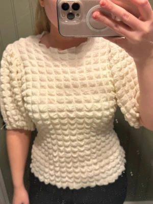 Vit puffärmad topp från Wera - Supersöt vit topp från Wera med puffärmar och ett unikt rutigt strukturmönster. Toppen har rund halsringning och är gjord i ett mjukt, stretchigt material som ger en skön passform. Perfekt för dig som vill sticka ut lite extra.