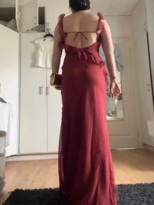 Balklänning - Säljer en vinröd långklänning med volangdetaljer, hög slits och öppen rygg med knytband. Klänningen har breda axelband och en elegant, flytande passform. Perfekt för dig som vill sticka ut med en romantisk look.