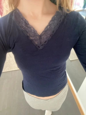 Mörkblå topp med spetsdetalj - Snygg mörkblå långärmad topp med v-ringning och spetsdetalj runt halsen. Figurnära passform som framhäver siluetten. Perfekt att matcha med både kjol och jeans för en trendig look.