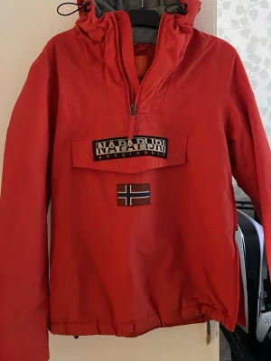 Orange/röd  vindjacka från Napapijri - Snygg röd vindjacka från Napapijri med stor magficka, halv dragkedja och norsk flagga på framsidan. Jackan har huva, tejpade sömmar och fleecefoder på insidan för extra komfort. Perfekt för blåsiga dagar och streetwear-stil.
