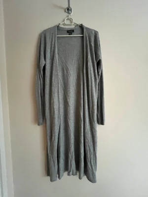 Lång grå kofta från H&M - Lång och öppen grå kofta från H&M med lång ärm och enkel design. Tillverkad i ett mjukt och tunt material som faller snyggt. Perfekt att slänga över en t-shirt eller topp för en avslappnad look.