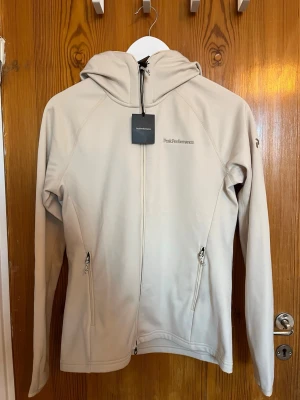 Beige hoodie från Peak Performance - Snygg beige hoodie från Peak Performance med dragkedja framtill och två fickor med dragkedja. Tillverkad i mjukt material och har en stilren logga på bröstet. Perfekt för dig som gillar enkel och clean stil. Huva med dragsko för extra komfort.