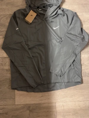 Nike vindjacka  - Snygg grå vindjacka från Nike med dragkedja framtill och huva. Jackan har en minimalistisk design med vit Nike-logga på bröstet och är tillverkad i lätt, vindtätt material. Perfekt för blåsiga dagar och enkel att matcha med olika outfits.