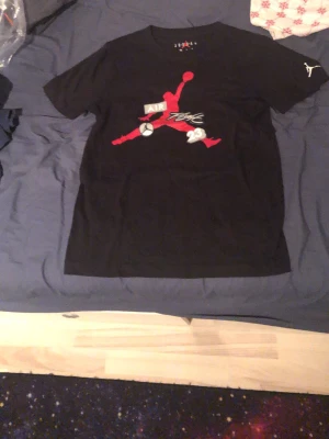 Svart Air Jordan t-shirt med tryck - Säljer en svart t-shirt från Jordan med det klassiska Jumpman-trycket i rött och vitt på bröstet. T-shirten har rund hals och korta ärmar, samt texten 'Air', 'Flight' och siffran 23 på framsidan. Perfekt för dig som gillar streetwear och sportig stil.