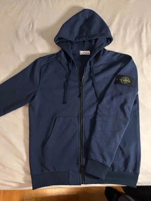 Blå hoodie från Stone Island - Säljer en blå hoodie från Stone Island med dragkedja framtill och klassisk patch på vänster ärm. Tillverkad i mjuk bomull för skön känsla och stilren look. Har endast använt ett fåtal gånger då armarna va lite för korta för mig. Står L men passar bättre på M