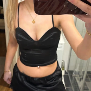 Svart croppad linnetopp - Snygg svart croppad linnetopp med tunna axelband och v-ringad urringning. Toppen har en figurnära passform och glansigt material som ger en trendig look. Perfekt att styla med kjol eller jeans för en cool outfit.