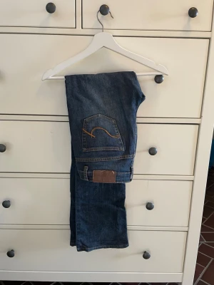 Blå bootcut jeans  - Säljer ett par klassiska blå bootcut jeans  med snygga kontrastsömmar och ikonisk bakficka med broderad detalj. Jeansen har normal midja och är tillverkade i slitstark denim, perfekta för en avslappnad och trendig look.