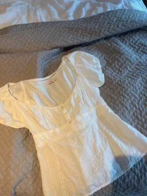 Vit blus med puffärm från Nelly - Söt vit blus från Nelly med puffiga korta ärmar och dekorativa knappar framtill. Blusen har en lätt rynkad struktur, rundad urringning och spetsdetaljer längs kanterna. Perfekt för en trendig och feminin look.