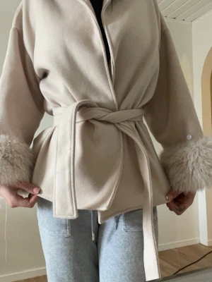 Beige kappa med fuskpälsdetaljer men känns som riktig päls - Stilren beige kappa med knytskärp i midjan och breda ärmslut dekorerade med fluffig fuskpäls. Kappan har en avslappnad passform och mjukt material som ger en lyxig känsla. Perfekt för dig som vill ha en trendig och elegant look. Den är helt ny säljer då den e lite för liten