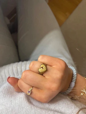 Krossat hjärta ring - Säljer en chunky ring i guldfärg med en unik, nästan skulptural form. Ringen har en matt finish och är tillverkad i metall. Perfekt för dig som gillar statement-smycken och vill sticka ut med din stil.