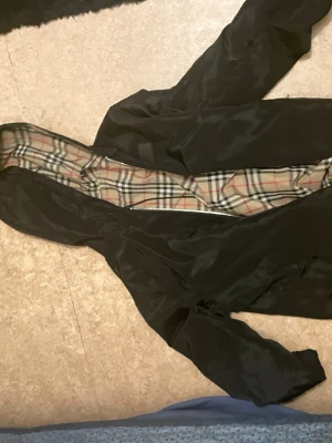 Svart vindjacka från Burberry - Svart vindjacka från Burberry med klassiskt rutigt foder i beige, rött och svart. Jackan har dragkedja framtill och huva. Materialet är glansigt och lätt, perfekt för blåsiga dagar. Ikonisk Burberry-label på insidan. Snygg och tidlös design.de är samma jacka som sticky använd i sviten låten jag säljer den ba för jag vill inte ha den längre 