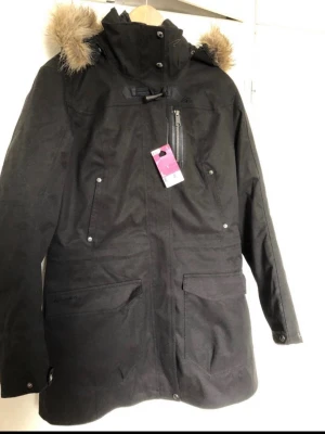 Svart parkasjacka från Quechua - Svart parkasjacka från Quechua med avtagbar huva och fuskpälsdetalj. Jackan har flera praktiska fickor med dragkedja och tryckknappar, hög krage och justerbar dragsko vid halsen. Perfekt för kalla dagar och riktigt snygg till vintern.