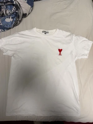 Vit t-shirt från Ami Paris med hjärtdetalj - Säljer en stilren vit t-shirt från Ami Paris med det ikoniska röda hjärtat och bokstaven A broderat på bröstet. Klassisk passform och rund halsringning. Perfekt till jeans eller shorts för en clean look. Storleken är L men passar även till stora m