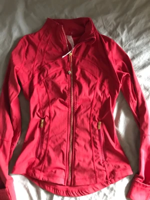 Röd träningsjacka från Lululemon - Snygg röd träningsjacka från Lululemon med figurnära passform och hel dragkedja framtill. Jackan har två fickor med guldfärgade dragkedjor och hög krage. Tillverkad i ett stretchigt funktionsmaterial som passar perfekt till träning eller löpning.