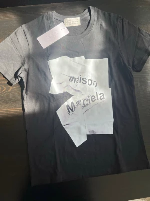 Maison Margiela svart t-shirt - Snygg svart t-shirt från Maison Margiela med ett stort vitt grafiskt tryck framtill. Klassisk rund hals och korta ärmar. Tillverkad i mjuk bomull för skön känsla. Perfekt för dig som gillar statement-plagg och modern design.