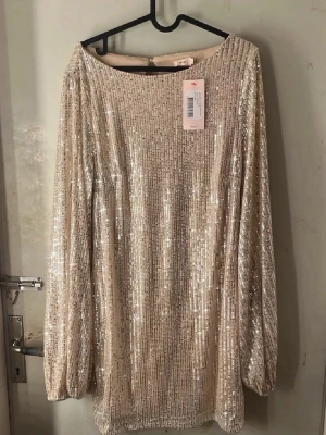 Glittrig kortklänning med paljetter - Supersnygg kortklänning från Nelly  i champagnefärgad ton, täckt av glittriga paljetter. Klänningen har långa, puffiga ärmar och en öppen ryggdetalj. Perfekt för dig som vill sticka ut med en festlig look. Rak passform och rund halsringning.