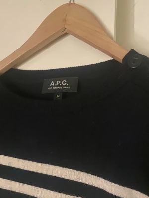 Randig stickad tröja från A.P.C. - Säljer en svart och vit randig stickad tröja från A.P.C. Tröjan har rund halsringning med knappdetalj på axeln och långa ärmar. Klassisk marin stil med smala ränder och ribbade muddar. Perfekt för dig som gillar stilrena plagg.