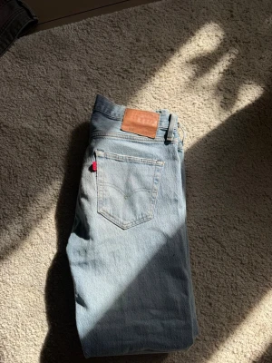 Levi's ljusblå raka jeans - Säljer ett par klassiska ljusblå jeans från Levi's med raka ben och femficksmodell. Jeansen har den ikoniska röda Levi's-lappen på bakfickan och en brun läderpatch i midjan. Tillverkade i slitstark denim, perfekta för en avslappnad stil. Säljer pga att de inte passar och är ej använda                                                     Hör av er vid funderingar!