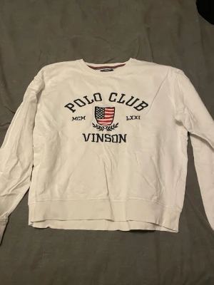 Vit tröja från Polo Club Vinson - Vit tröja med rund hals och tryck framtill med texten 'Polo Club Vinson' samt amerikansk flagga och romerska siffror. Perfekt för en avslappnad och sportig stil.