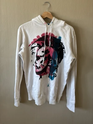 Vit hoodie med tryck från Diesel - Säljer en vit hoodie från Diesel med ett stort, färgstarkt ansiktstryck i rosa, svart och blått på framsidan. Hoodien har huva med dragsko och långa ärmar. Perfekt för dig som gillar streetwear och vill sticka ut med en unik design.