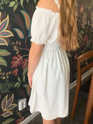 Vit offshoulder kortklänning - Söt vit kort klänning med offshoulder och puffärmar. Nyskick, endast använd en gång. Perfekt till skolavslutning. Storlek 152.