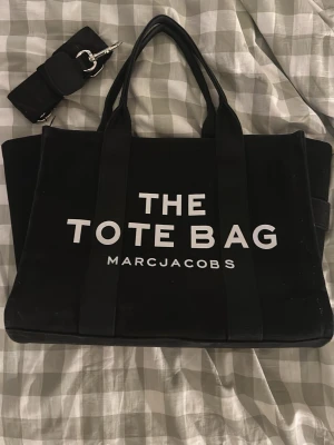 Svart shoppingväska Marc Jacobs - Snygg svart shoppingväska från Marc Jacobs med texten 'THE TOTE BAG' på framsidan och logotyp på baksidan. Väskan har två långa handtag och medföljer en avtagbar axelrem. Tillverkad i slitstarkt tygmaterial och har en stilren, boxig form.