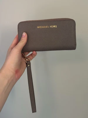 Brun plånbok från Michael Kors - Snygg brun plånbok från Michael Kors i tåligt saffianoläder. Plånboken har guldfärgade detaljer, dragkedja runtom och en avtagbar handledsrem. Klassisk och stilren design med logga i guld framtill. Lite slitet på ”flappen” och loggan (se bilder), annars är den i fint skick. 