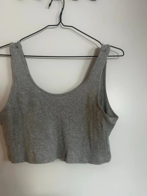 Grå ribbad croppad linnetopp - Snygg grå croppad linnetopp i ribbad bomullsmix. Toppen är ärmlös med breda axelband och rundad halsringning, perfekt för en avslappnad och trendig look. Enkel att matcha med jeans eller kjol. Rök och djurfritt hem. Pruta gärna 🌸