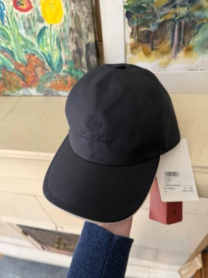 Loro Piana keps  - Snygg mörkblå keps från Loro Piana med diskret broderad logga framtill. Utsidan är slät och stilren, medan insidan har ett rutigt foder i beige, grönt och rött. Klassisk böjd skärm och justerbar passform. Perfekt accessoar för en clean och modern look.