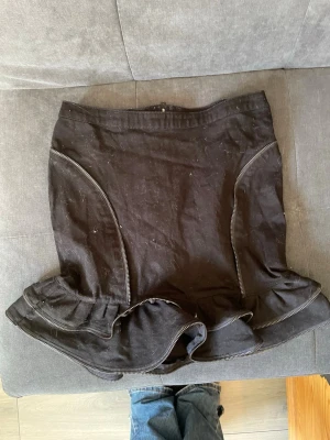 Svart volangkjol med dragkedjor - Säljer en svart kortkjol med volangkant och dekorativa dragkedjor längs sidorna. Kjolen har en markerad midja och en synlig dragkedja bak. Perfekt för dig som gillar edgy detaljer och vill sticka ut.