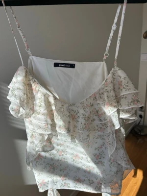 Blommig volangtop från Gina Tricot - Söt blommig topp från Gina Tricot med tunna justerbara axelband och volangdetaljer framtill. Toppen är vit med små rosa och gröna blommor, har en luftig passform och är perfekt för varma dagar. Materialet känns lätt och somrigt🥂💗