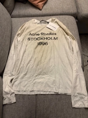 Vit långärmad topp Acne Studios - Vit långärmad topp från Acne Studios med tryck 'Acne Studios STOCKHOLM 1996' på bröstet. Tillverkad i mjuk bomull och har en avslappnad passform. Perfekt för en clean och stilren look.