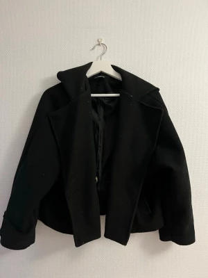 Svart kort ullrock från New collection  - Stilren svart kort ullrock från New collection med bred krage och dekorativa knappar vid ärmsluten. Rocken har en rak passform och är helfodrad för extra komfort. Perfekt för dig som vill ha en klassisk look med modern twist.