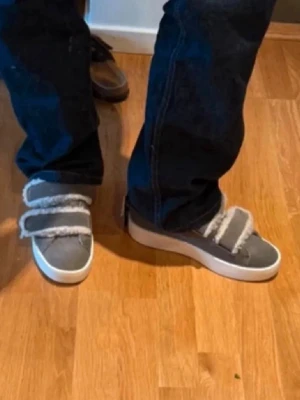 Grå sneakers med fuskpäls och kardborre - Unika sneakers i grått tyg med två breda kardborreband och fluffig fuskpälsdetalj. Skorna har vit sula och en avslappnad, chunky look som sticker ut. Perfekta för dig som vill ha något annorlunda och bekvämt. Använd endast 1 gång.