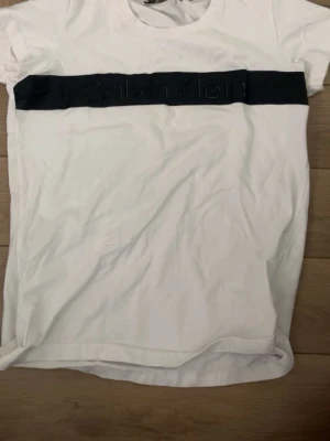 Vit Calvin Klein t-shirt med svart band - Vit t-shirt från Calvin Klein med ett svart band över bröstet där loggan är diskret broderad. Klassisk rund halsringning och korta ärmar.