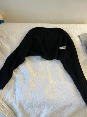 Svart ribbad bolero från Gina Tricot - Säljer en svart ribbad bolero från Gina Tricot. Den har lång ärm och är gjord i ett mjukt, stretchigt material. Perfekt att slänga över ett linne eller klänning för en cool lager-på-lager-look.