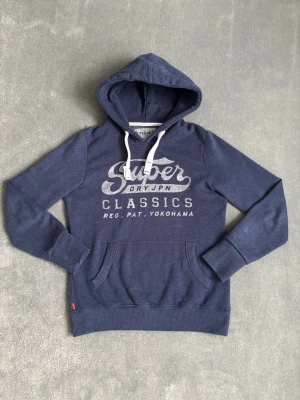 Marinblå Superdry Hoodie - Hoodie från Superdry, mycket bra skick. ”Superdry” loggan är inte sliten som den kanske ser ut på bilderna utan den är silvrig och lite glimrande. Storlek S. Hör av dig vid minsta fundering!😊